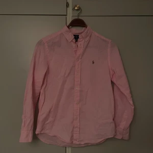 Ralph Lauren Skjorta - Riktigt fet rosa skjorta från Ralph Lauren! Klassisk button-down krage och broderad logga på bröstet. Skjortan har långa ärmar, knappar framtill och är gjord i mjuk bomull. Jag är 170 lång och väger 57 kg, den passar perfekt på mig! Perfekt för dig som gillar stilrena och fräscha plagg med ikonisk design.