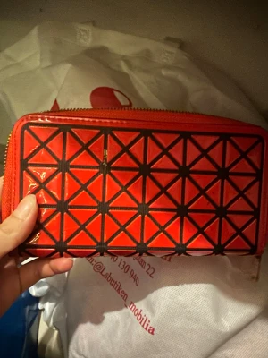 Röd geometrisk plånbok med dragkedja - Cool röd plånbok med svart geometriskt mönster på framsidan. Plånboken har en praktisk dragkedja runtom och flera fack för kort och mynt. Materialet är blankt syntet och insidan är röd med beige detaljer. Perfekt för dig som gillar grafisk stil och vill ha ordning på dina grejer.