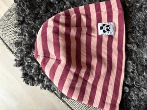 Randig mössa från Mini Rodini - Säljer en snygg mössa från Mini Rodini med breda ränder i vinrött och ljusrosa. Mössan är tillverkad i ekologisk bomull och har en vit patch med Mini Rodinis ikoniska panda-ansikte framtill. Perfekt för dig som gillar coola detaljer och streetstyle.