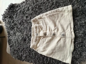 Beige manchesterkjol från Lindex Kids - Supersnygg naturvit kortkjol i manchester från Lindex Kids. Kjolen har raka fickor framtill och coola metallknappar med texten 'Love Every Day'. Perfekt för dig som gillar retrovibbar och vill ha en enkel men trendig look till ditt barn.