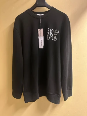 Svart sweatshirt från Palm Angels - Svart sweatshirt från Palm Angels med vit logotyp på bröstet och stor Palm Angels-text i vitt på ryggen. Tröjan har rund hals, långa ärmar och ribbade muddar. Perfekt för dig som gillar streetwear och vill sticka ut med en snygg designertröja.