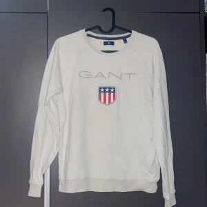 Vit sweatshirt från GANT med brodyr - Vit sweatshirt från GANT med broderad logga och sköld på bröstet. Tröjan har rund halsringning, långa ärmar och ribbade muddar. Klassisk och stilren design med inslag av rött och blått i brodyren. Perfekt för dig som gillar preppy stil. Jag har använt den typ 1-2 gånger och den kommer inte längre till användning 