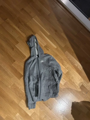 Grå hoodie från Nike med dragkedja - Säljer en grå hoodie från Nike för med vit logga på bröstet. I bra skick. Storlek 147-158 cm