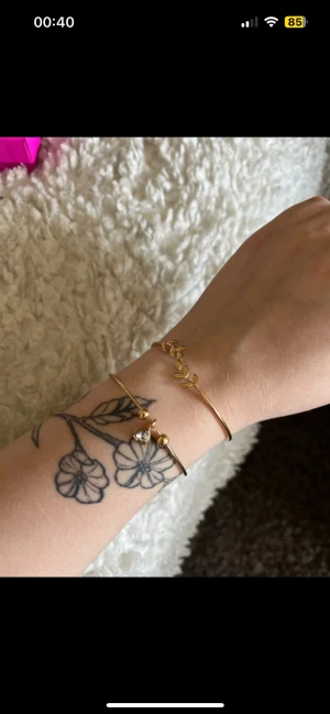Två guldiga armband med blad och sten - Säljer två tunna guldiga armband. Det ena har ett snyggt bladmotiv och det andra har små guldfärgade kulor och en glittrig sten. Perfekta för att ge din outfit lite extra shine och stil.