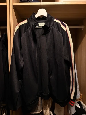 Svart bomberjacka från Gucci - Svart tracktop från Gucci med krage och dragkedja. Ärmarna har breda beige paneler med röda och marinblå ränder samt stor GUCCI-logga broderad längs ena ärmen. Jackan har en regular passform och sportig vibe, perfekt för dig som gillar streetstyle.