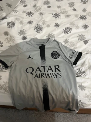 PSG Neymar Jr 10 grå fotbollströja - Snygg grå PSG fotbollströja med Neymar Jr och nummer 10 på ryggen. Framsidan har Jordan-logga, PSG-emblem och Qatar Airways tryck. Svart vertikal rand i mitten och svarta detaljer vid ärmslut. Tillverkad i lätt och ventilerande material.