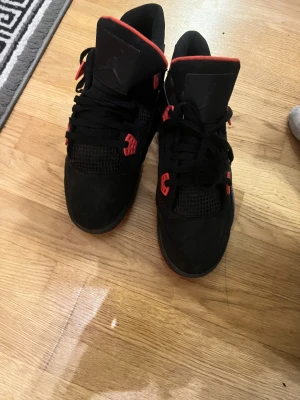 Svarta Nike Jordan 4 med röda detaljer - Nike Jordan 4 sneakers i svart med röda detaljer och meshpaneler. Skorna har snörning, platt sula och en klassisk siluett med högt skaft. Materialet är mestadels mocka och syntet, med en mjuk vadderad plös och röd insida. Perfekt för dig som gillar streetwear och ikoniska sneakers.