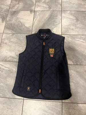 Mörkblå quiltad väst från Morris - Quiltad mörkblå väst från Morris med gul broderad logga och brittisk flagga på bröstet. Västen har ståkrage i manchester, dragkedja med läderdetalj och två sidofickor. Perfekt för dig som gillar klassisk stil med snygga detaljer.