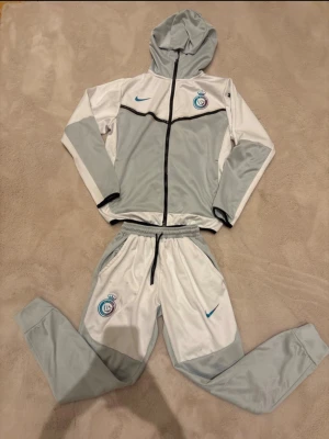 Nike ljusgrå och vit träningsset S - Nike träningsset i storlek S, med hoodie och matchande byxor. Setet är ljusgrått och vitt med svarta detaljer och har klubbmärke och Nike-logga i blått. Byxorna har snörning i midjan och muddar vid benslut. Materialet är 100% polyester, perfekt för sport och aktivitet.