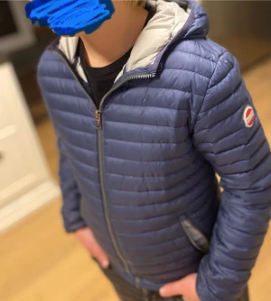 Mörkblå Colmar jacka - Snygg mörkblå pufferjacka från Colmar med huva och två dragkedjeförsedda fickor. Colmar-logga på ärmen. Perfekt för kyliga dagar och har en clean, look. Storleken är 154 i barn vilket är ungefär xs/xxs.