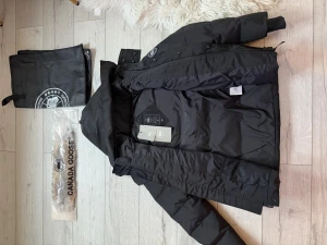 Canada Goose  - Säljer en svart dunjacka från Canada Goose med fluffig beige päls på huvan. Jackan har quiltad design, dragkedja och knappar framtill samt långärmade ärmar. Märkets logga syns på innerlappen och medföljande taggar. Perfekt för kalla dagar!