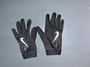 Nike Therma-Fit svarta träningshandskar - Säljer ett par svarta Nike Therma-Fit handskar med vit swoosh-logga på ovansidan. Handskarna är tunna, smidiga och har greppdetaljer i handflatan. Perfekta för löpning eller träning utomhus när det är kallt. Snygg, sportig look och bekväm passform.