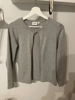 Grå kofta - En stilren grå kofta från Saint tropez i storlek XS. Koftan har rund halsringning, långärmade ärmar och stängs med en enkel knapp framtill. Materialet känns mjukt och är stickat, perfekt för lager på lager under kyliga dagar.
