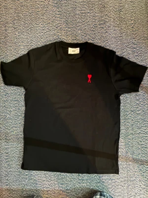 Svart t-shirt från Ami med rött hjärta - Svart t-shirt från Ami Alexandre Mattiussi med klassisk passform. På bröstet finns ett broderat rött hjärta med bokstaven A. T-shirten har rund halsringning och är tillverkad i mjuk bomull. Perfekt för en clean och stilren look.