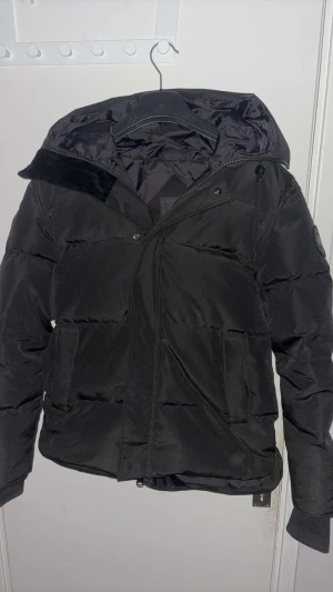 Svart pufferjacka från Canada Goose - Säljer en svart pufferjacka från Canada Goose med huva och klassisk rund logotyp på ärmen. Jackan har en clean look, breda quiltade sektioner och dragkedja framtill. Materialet är syntet och dun, perfekt för kalla dagar. Passformen är normal och jackan är långärmad.