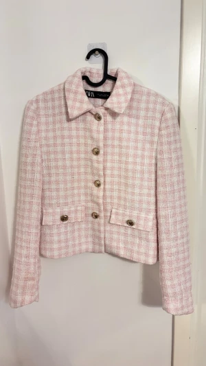 Rutig rosa kavajjacka från Zara - Superfin rutig kavajjacka från Zara i ljusrosa och vitt. Jackan är croppad med klassisk krage, snygga guldiga knappar och två fickor framtill. Materialet är strukturerat och känns lyxigt, perfekt för att lyfta vilken outfit som helst.