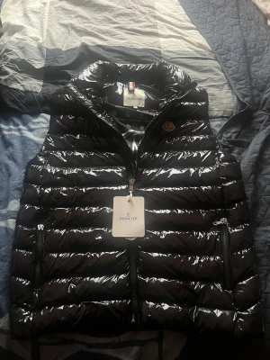Svart dunväst från Moncler - Säljer en svart, glansig dunväst från Moncler med quiltad design och hög krage. Västen har dragkedja framtill, två sidofickor med dragkedja och Moncler-logga på bröstet. Insidan har mönstrat foder med Moncler-tryck och en serietidningsetikett. Skick 9/10, lite strul med dragkedjan längst men de funkar. Storlek 4 vilket motsvarar strk L. Pris 1200kr. Vid snabb affär kan gå ner några hundra. Vid intresse eller frågor skriv!