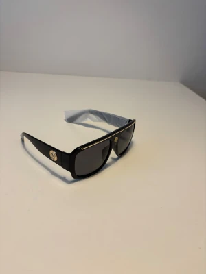 Versace liknande solglasögon  - Snygga svarta solglasögon från Versace med breda bågar och rektangulär form. De har mörka linser och guldiga emblem på sidorna samt en tunn guldfärgad detalj längs ovansidan. Perfekt accessoar för att lyfta din stil. Helt nya.