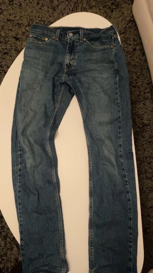 Levi's 505 blå jeans  - Klassiska blå Levi's 505 jeans med rak passform och fem fickor. Jeansen har en snygg mellanblå tvätt, traditionell knapp och dragkedja framtill samt läderpatch bak i midjan. Perfekta för en avslappnad och tidlös look. Storlek W29 L32. 