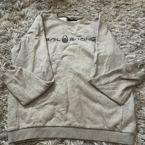 Grå Sail Racing sweatshirt - Snygg grå sweatshirt från Sail Racing med svart logotyp tryckt över bröstet. Tröjan har rund halsringning med ribbad kant och långa ärmar. Mjuk insida och klassisk passform, perfekt för chill dagar. Enkel och stilren design som funkar till jeans eller mjukis.