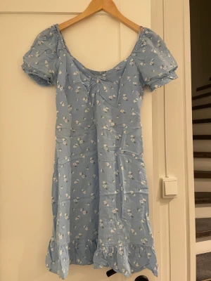 Blommig blå kortklänning Hollister - Supersöt ljusblå kortklänning från Hollister med vita blommor, puffärmar och volangkant nedtill. Smockad rygg och lätt v-ringad hals med rynkdetalj framtill. Perfekt för sommarens alla tillfällen, riktigt fräsch och ungdomlig vibe.