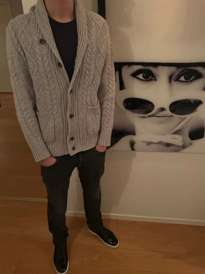 Grå stickad kofta med knappar - Säljer en grå stickad kofta med grovt kabelstickat mönster, stora knappar och två fickor framtill. Koftan har sjalkrage och mörka armbågslappar som snygg detalj. Perfekt för lager-på-lager under kyliga dagar.