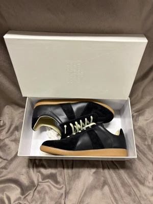 Maison Margiela svarta sneakers - Maison Margiela sneakers i svart skinn och mocka med kontrasterande vita snören och beige innersida. Snygg gummisula i naturfärg och klassisk rund tå. Ikonisk design med diskret logotyp på tungan, perfekt för dig som gillar stilrena och exklusiva sneakers.