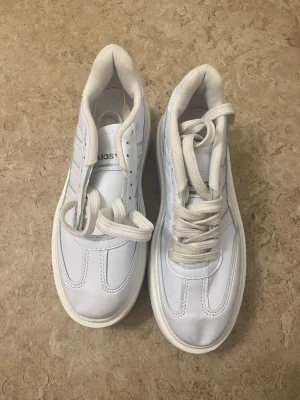 Vita Adidas sneakers med platt sula - Fräscha vita Adidas sneakers med klassiska tre ränder på sidan och logga bak på hälen. Skorna har platt sula, rund tå och snörning. Materialet är skinn med textilfoder och detaljer i gult och grönt på hälen och tungan. Perfekt för dig som gillar clean och sportig stil.