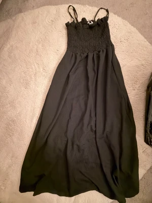 Svart långklänning från H&M - Svart långklänning från H&M med smockad överdel och tunna axelband. Klänningen har en flowy passform och är tillverkad i ett lätt tyg som ger en luftig känsla. Perfekt för varma dagar när du vill känna dig både bekväm och snygg.