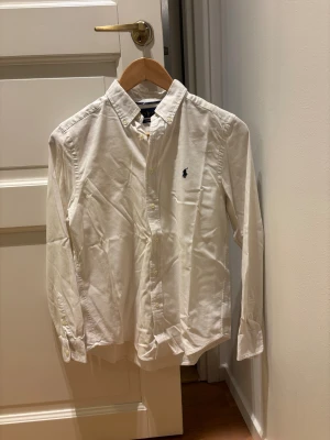 Vit skjorta från Ralph Lauren - Vit skjorta från Ralph Lauren med klassisk krage och knappar framtill. Broderad logga i marinblått på bröstet. Skjortan är långärmad och har en clean, stilren look. Tillverkad i bomull för skön känsla och bra passform. Kan även strykas vid köp 