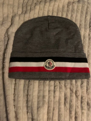  mössa - Snygg grå mössa från  med broderad logga framtill. Mössan har breda ränder i svart, vitt och rött längs kanten, vilket ger en cool kontrast. Tillverkad i mjuk stickad bomull som sitter skönt på huvudet. Perfekt för dig som gillar streetstyle och vill ha något som sticker ut.