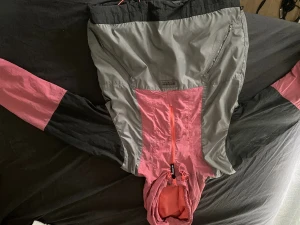 Adidas vindjacka XS i rosa och grå - Snygg vindjacka från Adidas i storlek XS. Jackan har färgblock i rosa, grått och svart, med huva och dragkedja framtill. Lätt och sportig design med praktiska fickor och justerbar huva. Perfekt för dig som gillar streetstyle och sportiga vibes.