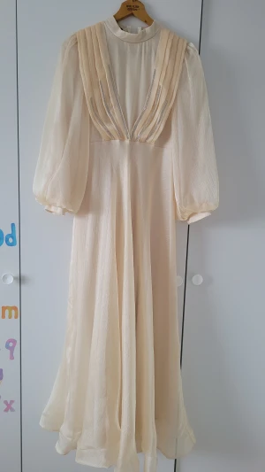 Champagnefärgad långklänning med glitter - Säljer en elegant långklänning i champagnefärg med puffiga långärmar och hög hals. Klänningen har glittriga detaljer framtill och är gjord i ett skimrande, lätt material som ger ett lyxigt intryck. Dragkedja i ryggen för enkel påklädning. 