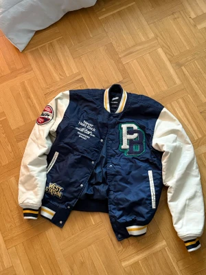 Blå och vit bomberjacka från Pull&Bear - Snygg bomberjacka från Pull&Bear i mörkblått med vita ärmar och broderade patches. Jackan har ribbade muddar med gula och vita ränder, tryckknappar och coola detaljer som stora bokstäver och tigrar på ryggen. Perfekt för dig som gillar streetstyle och collegevibe.