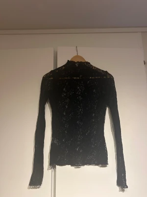 Svart spetsblus med lång ärm från MQ - Säljer en svart blus i genombruten spets med lång ärm och hög hals. Blusen har en slim passform och är helt täckt av vacker spets som ger en cool och elegant vibe. Perfekt att styla med jeans eller kjol för en trendig look.