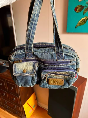 Blå denim handväska från Diesel - Säljer en blå handväska från Diesel i klassisk denim med flera fickor och detaljer i mässing. Väskan har två rejäla handtag, dragkedjor och en stor DIESEL-plakett framtill. Perfekt för dig som gillar street och retro vibes.