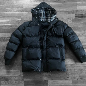 Svart pufferjacka med huva - Säljer en svart pufferjacka med huva och rutigt foder. Jackan har dragkedja och knappar framtill, två sidofickor med dragkedja och ribbade muddar vid ärmsluten. Perfekt för kalla dagar och riktigt skön att bära.