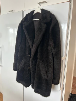 Svart  pälsjacka från Pimpernel - Säljer en fluffig och stilren mörkbrun pälsjacka från Pimpernel of Denmark. Jackan har bred krage, knappar framtill, fickor och ett bälte bak. Materialet är 65% acrilan och 35% modacrylic pile, vilket ger en mjuk och varm känsla. Perfekt för kalla dagar.