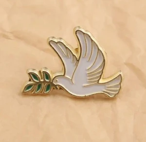 Fredsduva-pin med olivkvist - Superfin pin i form av en vit duva med guldkanter som håller en grön olivkvist i näbben. Perfekt accessoar att fästa på jackan eller väskan för att sprida fredsvibbar. Storlek ca 2,8 x 2 cm. Snygg detalj som sticker ut och passar alla stilar.