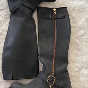 Svarta stövlar med dragkedja och spänne - Snygga svarta stövlar i skinn med dragkedja och dekorativt spänne. Perfekta för höst och vinter. Sulan har ett mönster för bra grepp. Stilrena och funktionella för alla tillfällen.