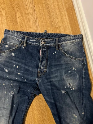 Dsquared2 blå jeans med slitningar - Säljer ett par blå jeans från Dsquared2 med coola slitningar och färgstänk för en riktigt edgy look. Klassisk femficksmodell med Dsquared2-detaljer på knapp och bakficka. Jeansen har rak passform och är tillverkade i robust denim. (Äkta)