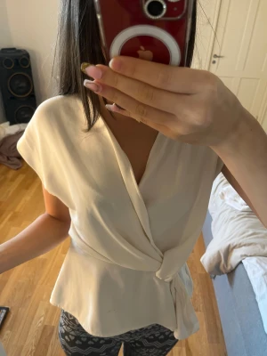 Krämvit omlottblus från Zara - Säljer en krämvit omlottblus från Zara i storlek S. Blusen har korta ärmar, v-ringning och knyt i midjan som ger en snygg siluett. Materialet är mjukt och har ett lätt fall, perfekt för dig som gillar stilrena och enkla plagg. Ett litet hål längst öpp på dragkedjan som ej märks av eller syns.