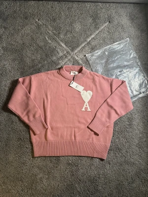 Rosa stickad tröja från Ami - Snygg rosa stickad tröja från Ami Alexandre Mattiussi med ribbad krage, muddar och nederkant. På bröstet finns ett stort vitt hjärta med bokstaven A. Perfekt för dig som gillar stilrena detaljer och vill ha en tröja med lite edge.