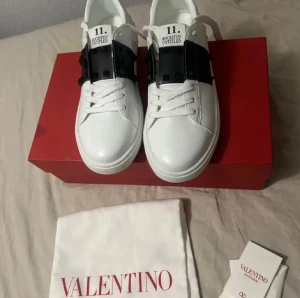 Valentino Rockstud Untitled dojor - Snygga vita Valentino Rockstud Untitled sneakers med svart band och ikoniska pyramidnitar över vristen. Skorna har rund tå, platt sula och snörning framtill. Tillverkade i skinn med coola detaljer både fram och bak. Perfekt för dig som gillar exklusiv streetstyle.