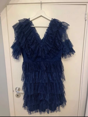 Marinblå spetsklänning från By Malina - Säljer en marinblå kortklänning från By Malina i storlek L men passar även M-S. Klänningen har flera lager med volanger i spets, v-ringad hals och korta ärmar med extra volangdetaljer. Den har en romantisk och trendig look som verkligen sticker ut.