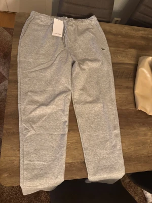 Grå mjukisbyxor från Lacoste L - Grå mjukisbyxor från Lacoste i klassisk fit med elastisk midja och snörning. Byxorna har sidofickor, en bakficka och den ikoniska krokodilloggan broderad på benet. Perfekta för chill eller träning, tillverkade i mjukt bomullsmaterial.