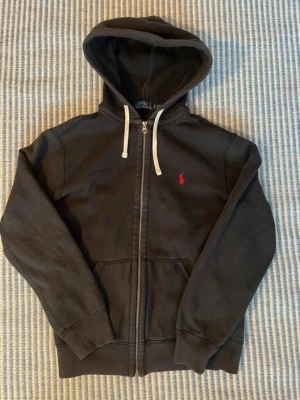 Svart hoodie från Polo Ralph Lauren - Svart hoodie från Polo Ralph Lauren med röd broderad logga på bröstet. Tröjan har huva med vita snören, dragkedja framtill och två fickor. Materialet är mjuk bomull och insidan har en skön, lite fleece-liknande känsla. Perfekt för chill dagar och streetwear.