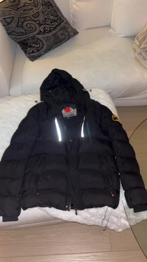 Svart pufferjacka från Zavetti Canada - Svart pufferjacka från Zavetti Canada med huva och reflekterande detaljer på bröstet. Jackan har broderad patch på ärmen, dragkedjor på fickorna och justerbar huva. Perfekt för kalla dagar och har en schysst streetstil. Kan lägga till fluff på huvan om man vill.