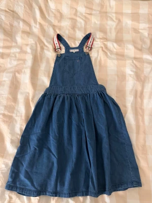 Jeansklänning Tommy Hilfiger 152 - Snygg jeansklänning från Tommy Hilfiger med klassiska logoband på axelbanden och en ficka framtill. Klänningen har en utställd kjol och coola detaljer i metall. Perfekt för dig som gillar streetstyle och denim.