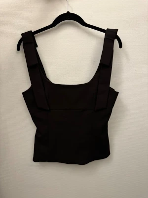 Svart topp med rosetter vid axlarna - Snygg svart korsett-topp med breda axelband och fyrkantig halsringning. Toppen har en stilren och strukturerad form som ger en cool siluett. Perfekt att styla med jeans eller kjol för en trendig look.